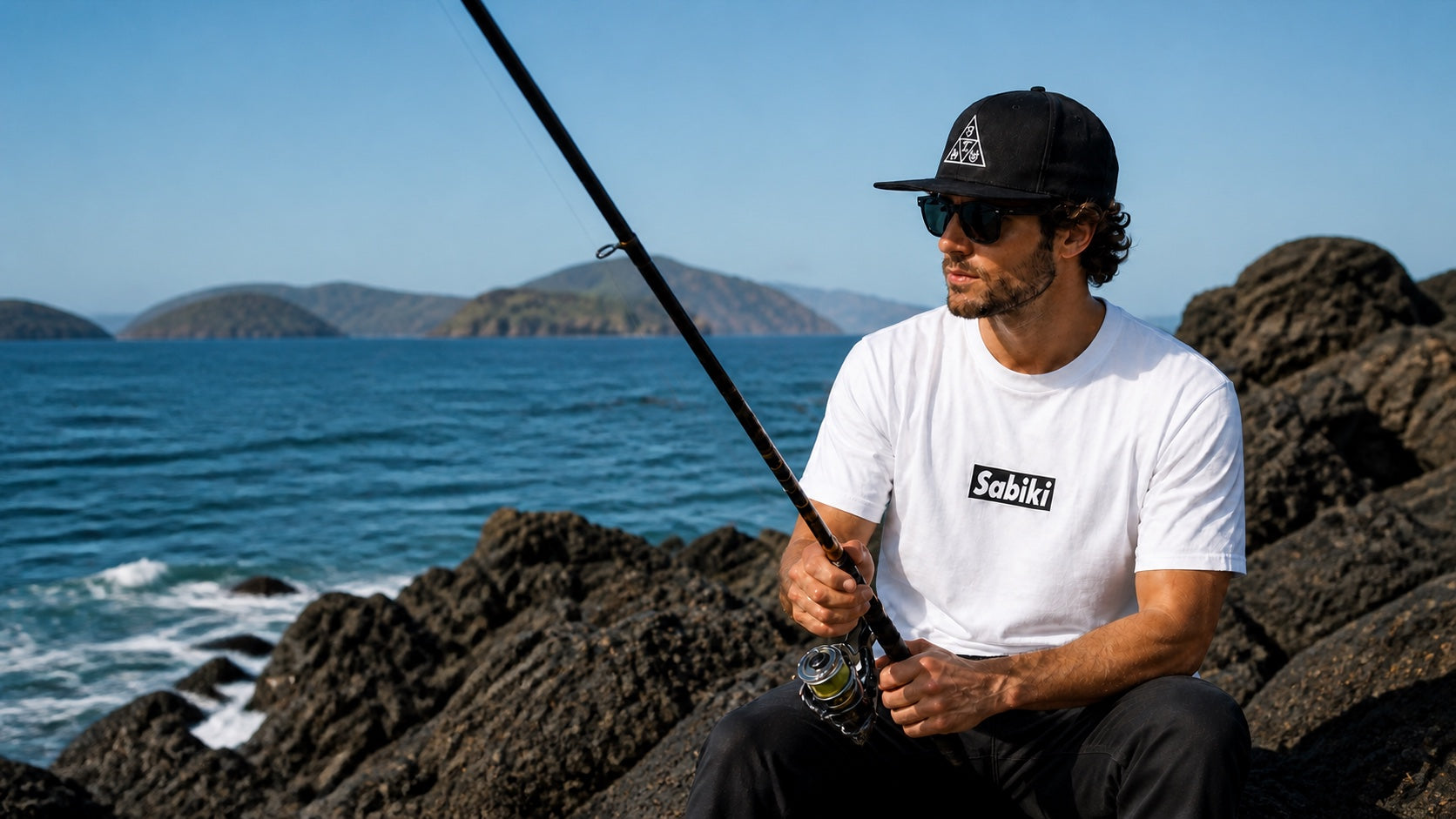 海辺でISUの白いTシャツと黒いDeltaキャップを着用し、釣り竿を持つアングラー